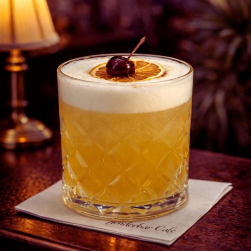 Amaretto Sour koktel sa penom od belanaca, serviran u Nick & Nora čaši sa sušenim kolutom limuna, u toplom barskom ambijentu.